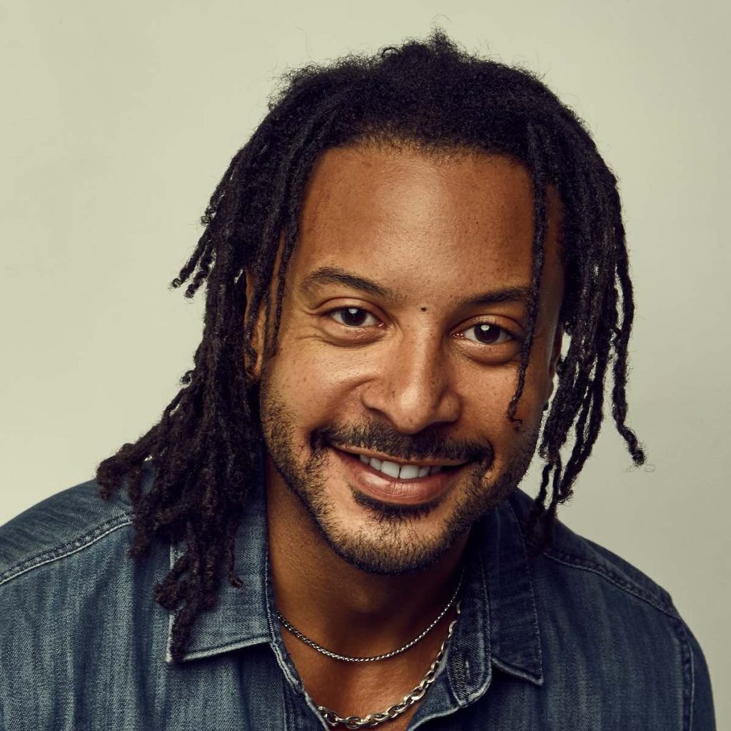A medium close headshot of a man in a denim shirt looking at the camera with an open-mouth smile