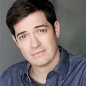 A close headshot of a man in a blue shirt looking at the camera with a slight closed-mouth smile