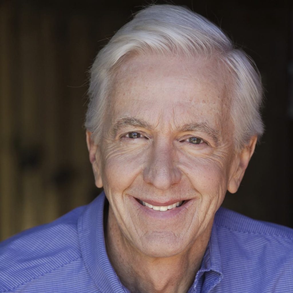 A close headshot of a man in a blue shirt looking at the camera with an open-mouth smile