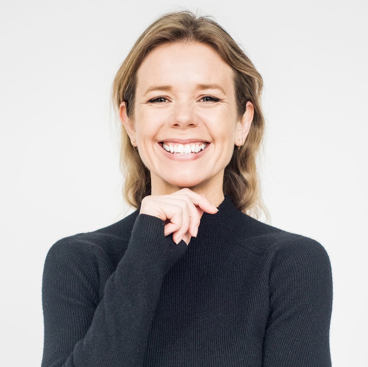 A medium of a woman in a black long-sleeved sweater, smiling broadly at the camera with one hand under her chin
