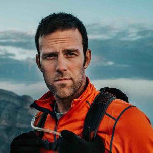 A medium headshot of a man in a orange parka and black knapsack looks at the camera with mountain and sky behind him