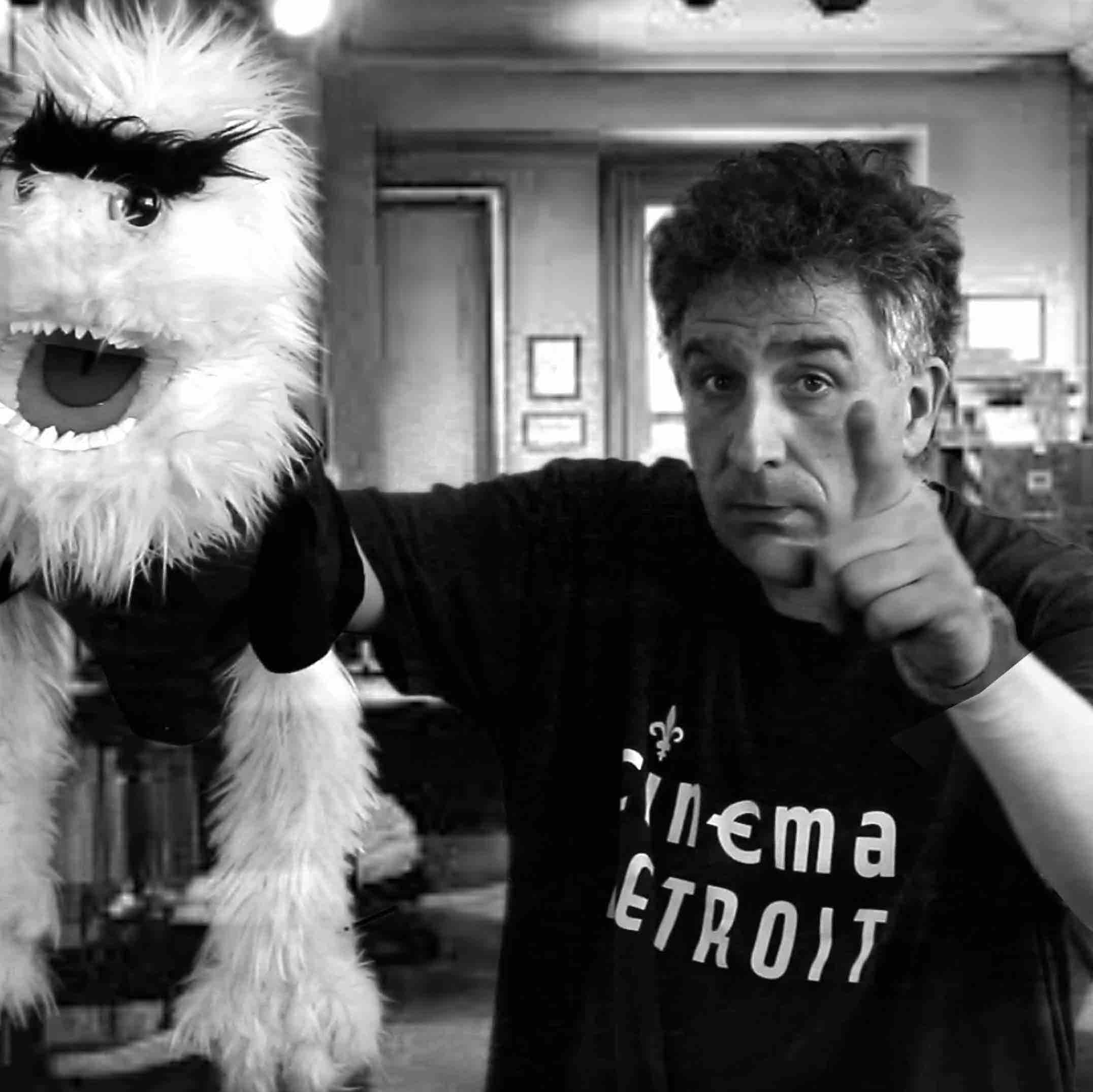 A medium black & white shot of a man pointing a finger at camera while holding a large fuzzy puppet in the other hand