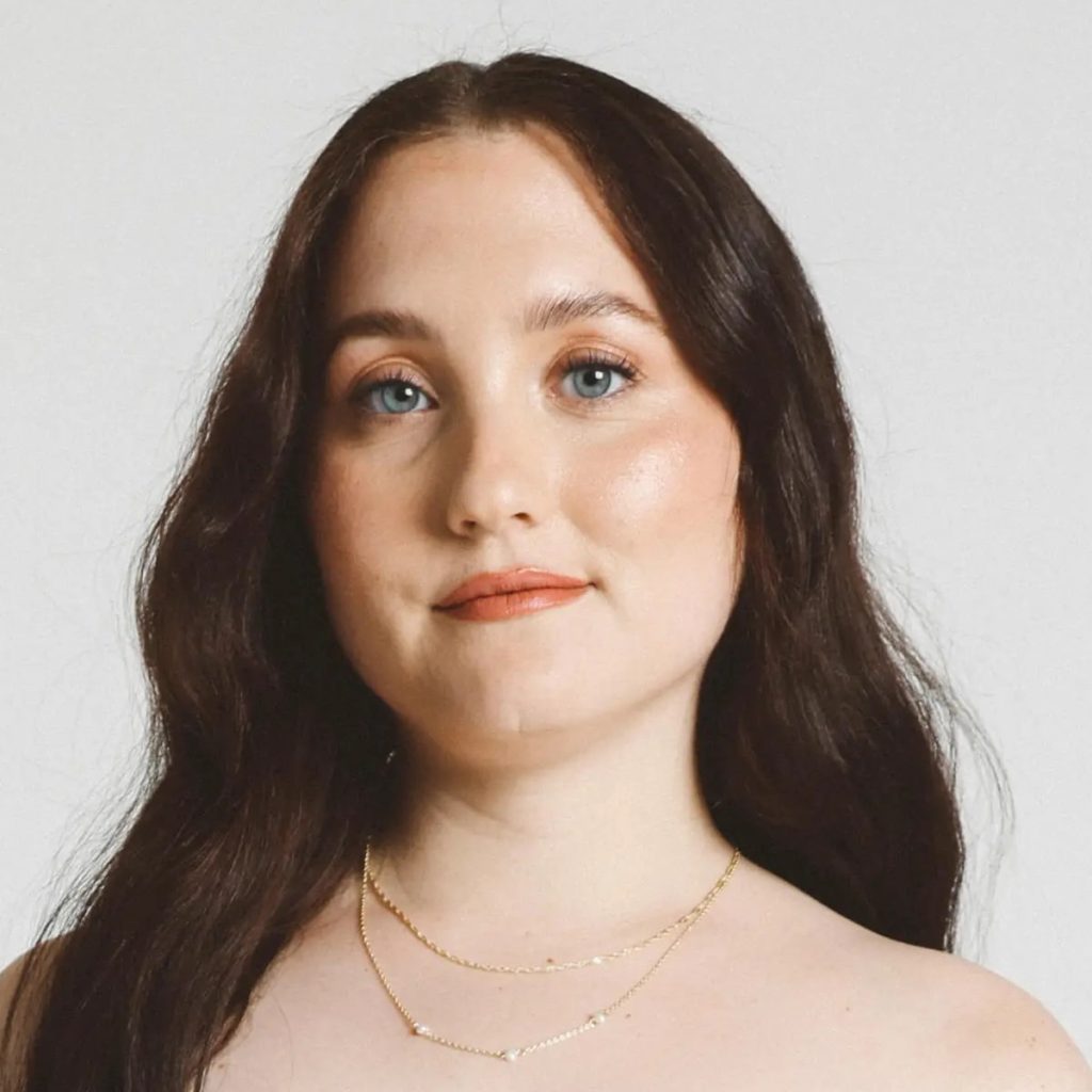 Young woman with long dark brown hair and blue eyes, wearing layered gold necklaces, looking directly at the camera against a pale background.
