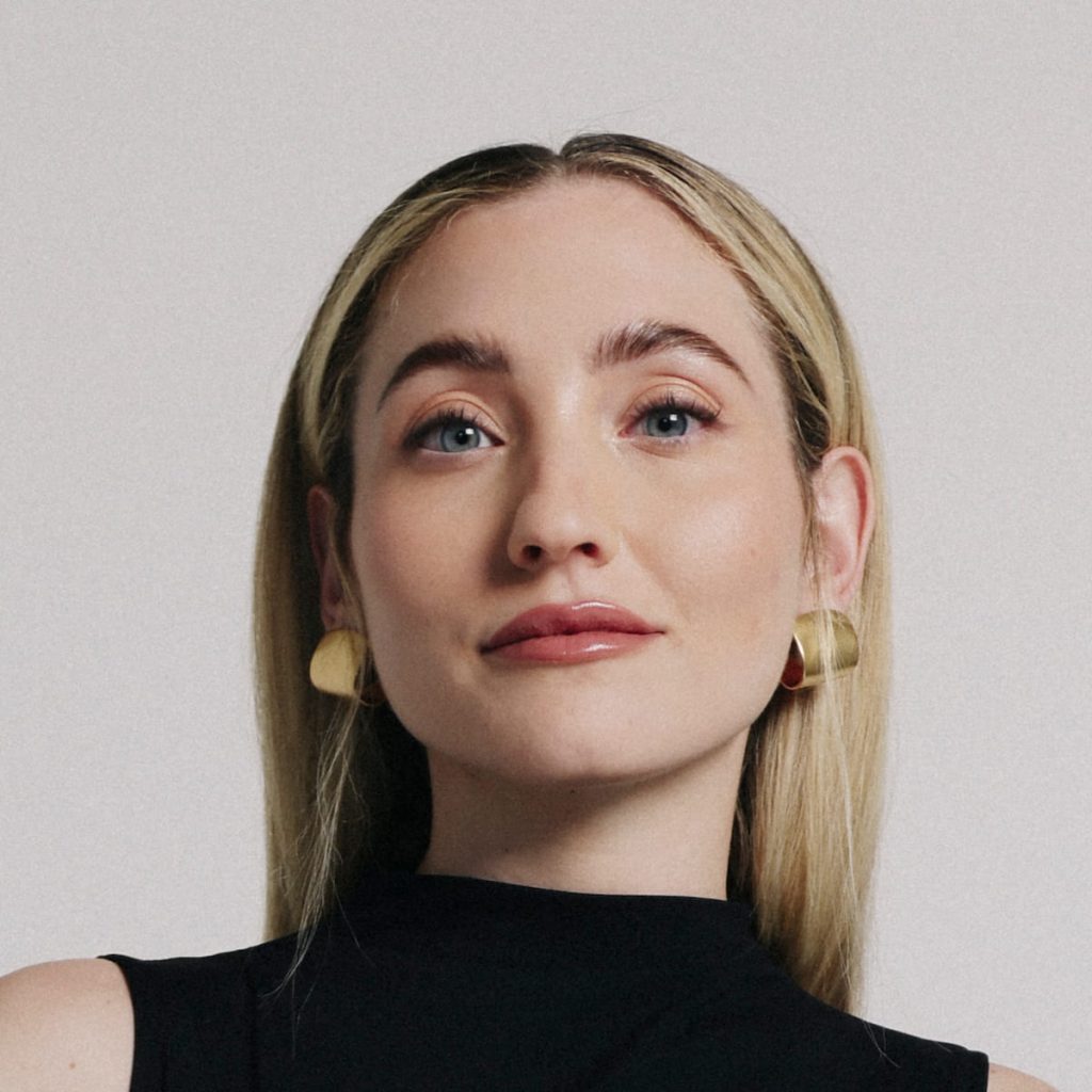 Headshot of a blonde woman in a black sleeveless top and gold earrings, smiling softly